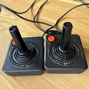 May include: Two black video game joysticks with square bases, a central vertical stick, and a single red circular button on the top left corner of each unit. Each joystick features a black ribbed circular base and includes a thin black wired cable.