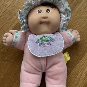 May include: A soft-bodied doll with a round face, dark hair, and a light skin tone. The doll wears a pink one-piece outfit, a ruffled bonnet with a colorful pattern, and a lavender bib featuring the embroidered text Cabbage Patch Kids BABYLAND.