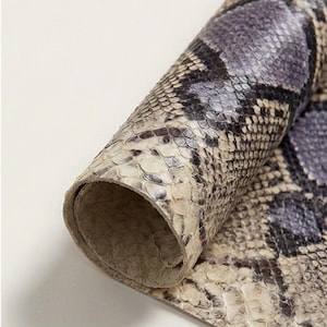 Puede incluir: Piel de serpiente enrollada con un patrón natural. El cuero muestra una mezcla de tonos grises, negros y crema, con una textura escamosa. El material está enrollado, revelando el reverso y el patrón detallado.
