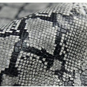 Peut inclure: Gros plan sur une matière texturée avec un motif peau de serpent. Le motif présente un mélange d'écailles grises, noires et blanches, créant un aspect réaliste de peau de reptile. La matière est légèrement incurvée.