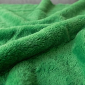 Può includere: Primo piano di una coperta verde brillante e soffice. Il tessuto ha una consistenza morbida e pelosa con pieghe e ombre visibili, creando una sensazione di profondità. La coperta sembra essere realizzata in ecopelliccia, perfetta per il calore.