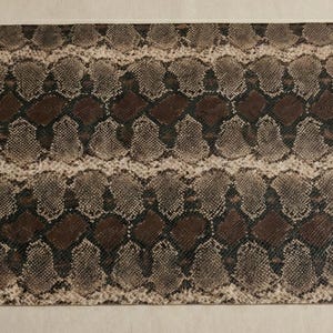Pode incluir: Uma peça retangular de material com um padrão de pele de cobra. O design apresenta um padrão repetitivo de escamas marrons, cinzas e bege. O material parece plano e pode ser usado para vários projetos de artesanato.