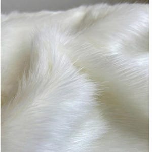 May include: Close-up of a white faux fur textile. The soft, fluffy material has long, flowing fibers, creating a textured surface. The lighting highlights the sheen and depth of the fur, suggesting a luxurious feel. The overall impression is one of warmth and comfort.