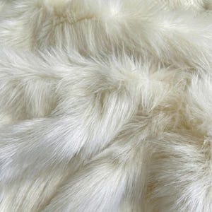 May include: Close-up of a cream-colored faux fur fabric. The fabric has long, soft, and fluffy fibers, creating a textured surface. The fibers are layered, with varying lengths and directions, giving the fabric a luxurious appearance.