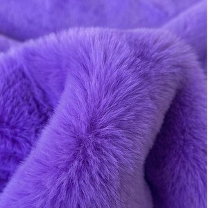 May include: Close-up of a vibrant purple faux fur fabric. The plush material has a dense, textured appearance with long, soft fibers. The color is a rich, saturated shade of purple, creating a luxurious and inviting look. The fabric is likely used for clothing or home decor.