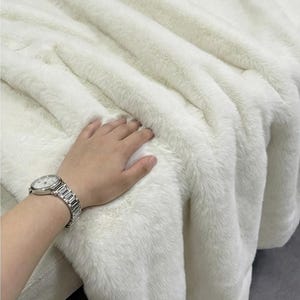 May include: A plush, white faux fur blanket with a soft, textured surface. The blanket is folded, creating deep ridges and shadows. The image shows a close-up of the blanket's texture, highlighting its luxurious appearance. A silver watch is visible.