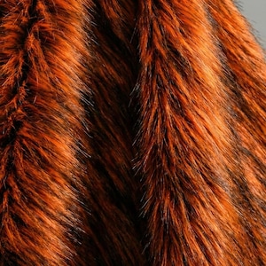 Może przedstawiać: Zbliżenie na teksturowaną, pomarańczowo-czarną tkaninę ze sztucznego futra. Tkanina ma długie, szorstkie włókna z mieszanką odcieni pomarańczowego i czarnego, tworząc wzór w paski. Materiał wydaje się miękki i luksusowy, odpowiedni do różnych projektów rzemieślniczych.