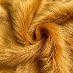 May include: Close-up of golden-yellow faux fur fabric. The plush material has long, flowing fibres with a slight sheen, creating a soft, textured appearance. The fibres swirl in a circular pattern.
