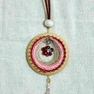 以下が含まれることがあります： ピンクと赤の糸で包まれた木製のフープが特徴の手作りのドリームキャッチャー。小さな赤いチャーム、ビーズのアクセント、紫色の羽のぶら下がりストランドが付いています。茶色の革紐から吊り下げられています。