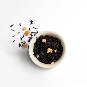 May include: A top-down view of a small, off-white bowl filled with dark tea leaves and small, light brown sugar cubes. Loose tea leaves and sugar cubes are scattered around the bowl on a white background. The tea leaves are a deep, rich brown color.