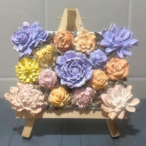 May include: A square canvas display featuring a collection of three-dimensional floral sculptures in shades of lavender, yellow, peach, and cream, arranged over a base of small grey pebbles, all supported by a light-coloured wooden tabletop easel.