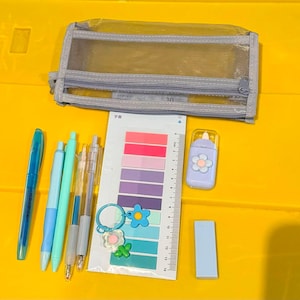 May include: A translucent grey mesh pencil case, a blue pen, a light blue pen, a mint green pen, two clear mechanical pencils, a sheet of adhesive sticky tabs with a ruler, a light purple correction tape, and a light blue rectangular eraser.