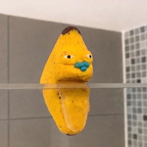 Könnte beinhalten: Eine gelbe, bananenförmige Figur mit brauner Spitze, kleinen schwarzen Augen und leuchtend blauen Lippen, die so positioniert ist, dass sie auf einer dünnen horizontalen Kante zu ruhen scheint und eine Spiegelung unter der Linie erzeugt.