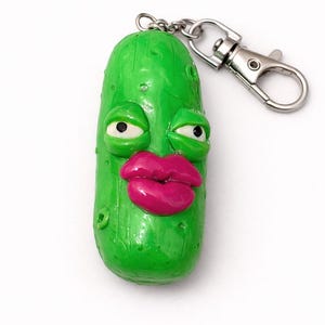 May include: A novelty keychain in the shape of a green pickle with a cartoon face. It features white eyes, a pink mouth, and a silver clasp. The keychain is designed to be attached to keys or bags.