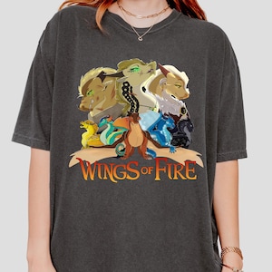 May include: Dark grey t-shirt with a vibrant illustration of dragon-like creatures. The design includes the text "WINGS OF FIRE" in orange and yellow. The shirt has short sleeves and a crew neck.