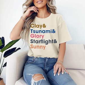 May include: A cream-coloured t-shirt with the words "Clay & Tsunami & Glory Starflight & Sunny" printed in various colours. The shirt is worn with blue jeans. The model is sitting on a white sofa.