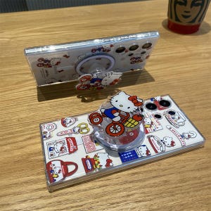 May include: Clear phone case with a Hello Kitty design. The case features a pop-up grip with a Hello Kitty character riding a bicycle. The case is decorated with various Hello Kitty-themed illustrations and text.