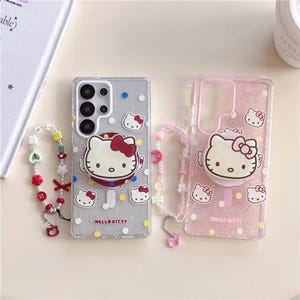 Pode incluir: Duas capas de telemóvel com designs Hello Kitty. Uma capa é transparente com glitter prateado e um pop-up Hello Kitty, a outra é rosa com glitter e um pop-up Hello Kitty. Ambas as capas têm a inscrição "Hello Kitty". Duas pulseiras de contas também estão incluídas.