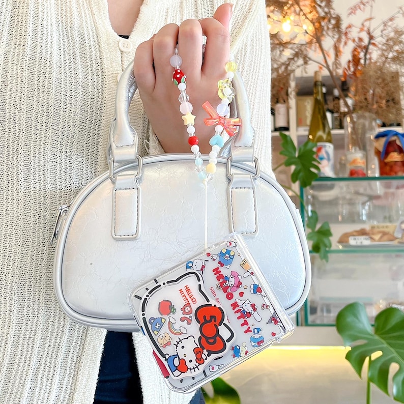 May include: A small, white handbag with silver-tone hardware and a beaded charm. A clear phone case with Hello Kitty characters is attached. The charm features colourful beads and a small bow.