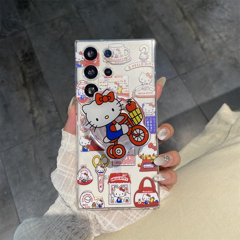 May include: Clear phone case featuring Hello Kitty designs. The case has a pop-up grip with a Hello Kitty graphic riding a tricycle. Additional Hello Kitty stickers and illustrations are scattered across the case, including a teacup, scissors, and a shopping bag.