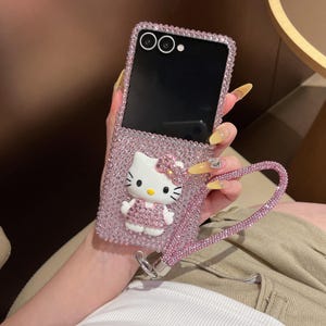 Puede incluir: Funda de tel&eacute;fono rosa con brillantes, con un dise&ntilde;o de Hello Kitty. La funda tiene una correa de mu&ntilde;eca a juego con brillantes y una figura 3D de Hello Kitty. La pantalla del tel&eacute;fono es negra. La funda est&aacute; dise&ntilde;ada para un tel&eacute;fono plegable.
