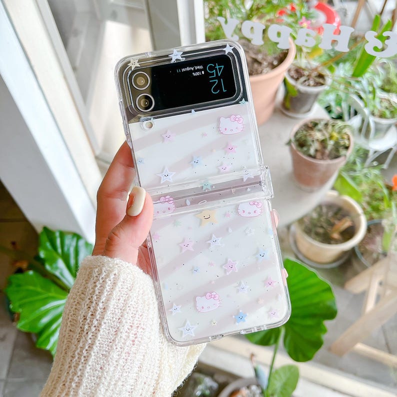 May include: Clear phone case with a white and pink striped pattern, featuring cartoon stars and Hello Kitty characters. The case is designed for a folding phone and has a screen displaying the time.