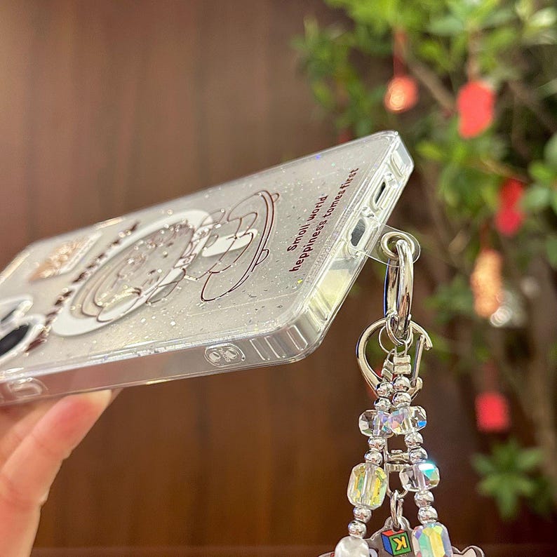 Puede incluir: Funda de tel&eacute;fono transparente con un dise&ntilde;o brillante. La funda presenta un personaje de dibujos animados y el texto "Small world, happiness comes first". Un charm plateado con cuentas y la letra "K" cuelga de la funda.