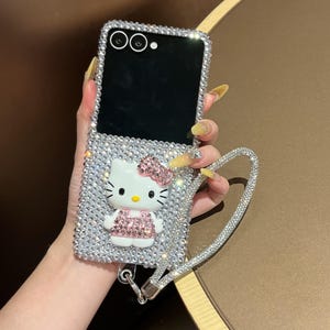 Puede incluir: Funda de tel&eacute;fono plateada cubierta de pedrer&iacute;a con un dise&ntilde;o de Hello Kitty con lazo y vestido rosa. La funda tiene una correa de mu&ntilde;eca de pedrer&iacute;a a juego. La funda est&aacute; dise&ntilde;ada para un tel&eacute;fono plegable.