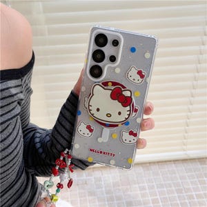 K&ouml;nnte beinhalten: Transparente Handyh&uuml;lle mit Hello Kitty-Design. Die H&uuml;lle verf&uuml;gt &uuml;ber einen Pop-up-Griff mit einem Hello Kitty-Gesicht und mehreren Hello Kitty-Gesichtsgrafiken. Die H&uuml;lle hat einen glitzernden Hintergrund mit blauen, gelben und wei&szlig;en Punkten. Die Worte "HELLO KITTY" sind auf der H&uuml;lle aufgedruckt.