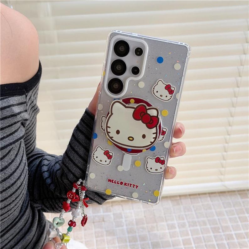 K&ouml;nnte beinhalten: Transparente Handyh&uuml;lle mit Hello Kitty-Design. Die H&uuml;lle verf&uuml;gt &uuml;ber einen Pop-up-Griff mit einem Hello Kitty-Gesicht und mehreren Hello Kitty-Gesichtsgrafiken. Die H&uuml;lle hat einen glitzernden Hintergrund mit blauen, gelben und wei&szlig;en Punkten. Die Worte "HELLO KITTY" sind auf der H&uuml;lle aufgedruckt.