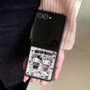 May include: Black folding smartphone with a Hello Kitty design. The case features two Hello Kitty characters in black and white outfits, with the text "HELLO KITTY" above them. The background is a marble pattern with small hearts.