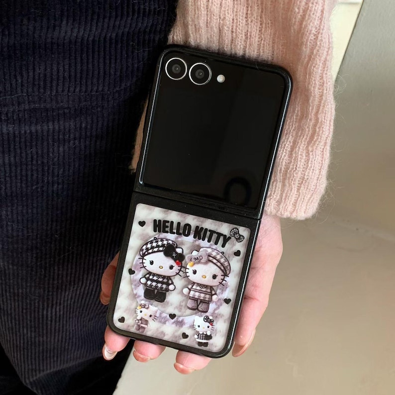 May include: Black folding smartphone with a Hello Kitty design. The case features two Hello Kitty characters in black and white outfits, with the text "HELLO KITTY" above them. The background is a marble pattern with small hearts.