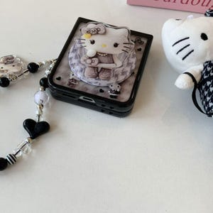 May include: Black phone case with a Hello Kitty design, featuring a clear pop socket with a Hello Kitty character. The case has a marble pattern and small heart details. A beaded phone strap with black heart and bead accents is also visible.