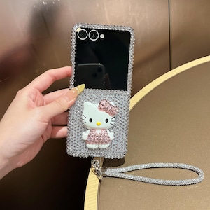 Puede incluir: Una funda plateada para tel&eacute;fono plegable cubierta de pedrer&iacute;a con un dise&ntilde;o de Hello Kitty. La funda tiene una correa para la mu&ntilde;eca y una pantalla negra. La figura de Hello Kitty tiene un lazo y un vestido rosas.