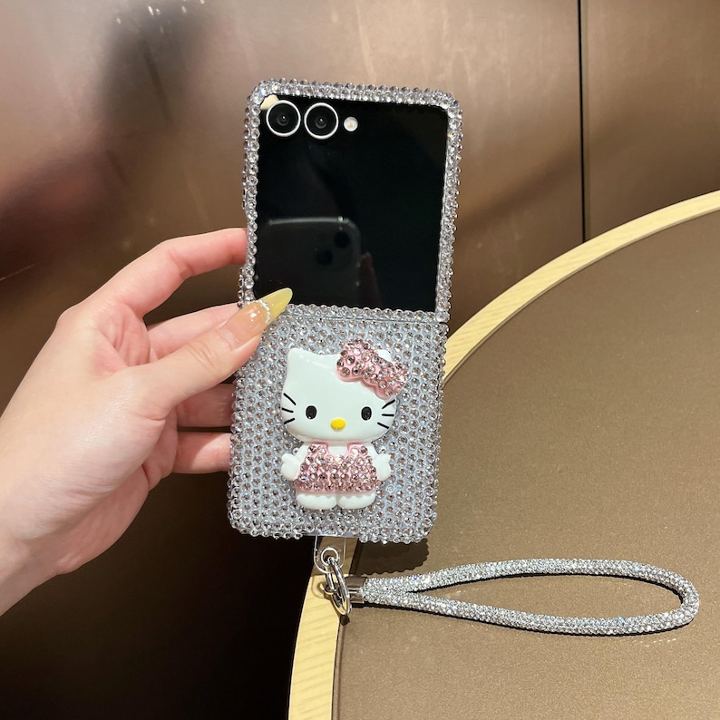 Puede incluir: Una funda plateada para tel&eacute;fono plegable cubierta de pedrer&iacute;a con un dise&ntilde;o de Hello Kitty. La funda tiene una correa para la mu&ntilde;eca y una pantalla negra. La figura de Hello Kitty tiene un lazo y un vestido rosas.