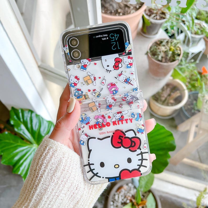 May include: Clear phone case with a Hello Kitty design. The case has a white background with various Hello Kitty illustrations and the text "Hello Kitty 50th Anniversary". The case is designed to fold open.