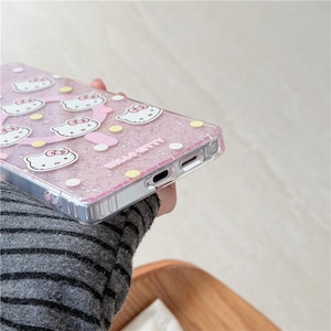 K&ouml;nnte beinhalten: Transparente Handyh&uuml;lle mit pinkfarbenem Glitzerhintergrund und Hello Kitty-Grafiken. Die H&uuml;lle hat einen silbernen Rahmen und zeigt wei&szlig;e Hello Kitty-Gesichter, rosa Schleifen sowie gelbe und wei&szlig;e Punkte. Der Text "HELLO KITTY" ist sichtbar.