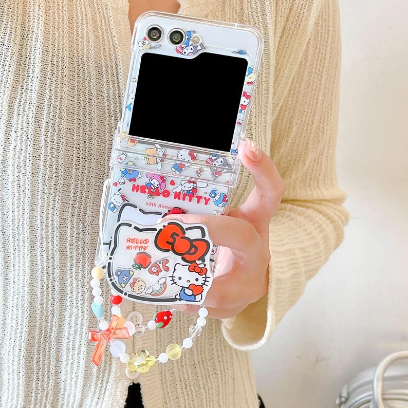 May include: Clear phone case with a Hello Kitty design. The case has a flip design and a wrist strap with beads and charms. The case features a Hello Kitty bow and the words "Hello Kitty 50th Anniversary".