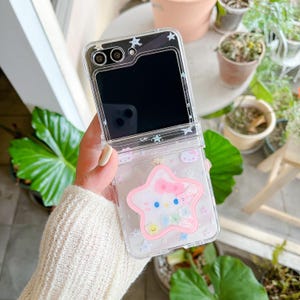 May include: Clear phone case with a pink star-shaped design featuring a cartoon character with a bow. The case has a flip-open design and is decorated with small stars and other cartoon characters.