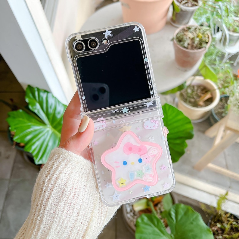May include: Clear phone case with a pink star-shaped design featuring a cartoon character with a bow. The case has a flip-open design and is decorated with small stars and other cartoon characters.