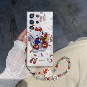 May include: Clear phone case with a Hello Kitty design. The case features a pop-up grip with a Hello Kitty graphic riding a bicycle. A matching beaded wrist strap with red and white accents is also included.