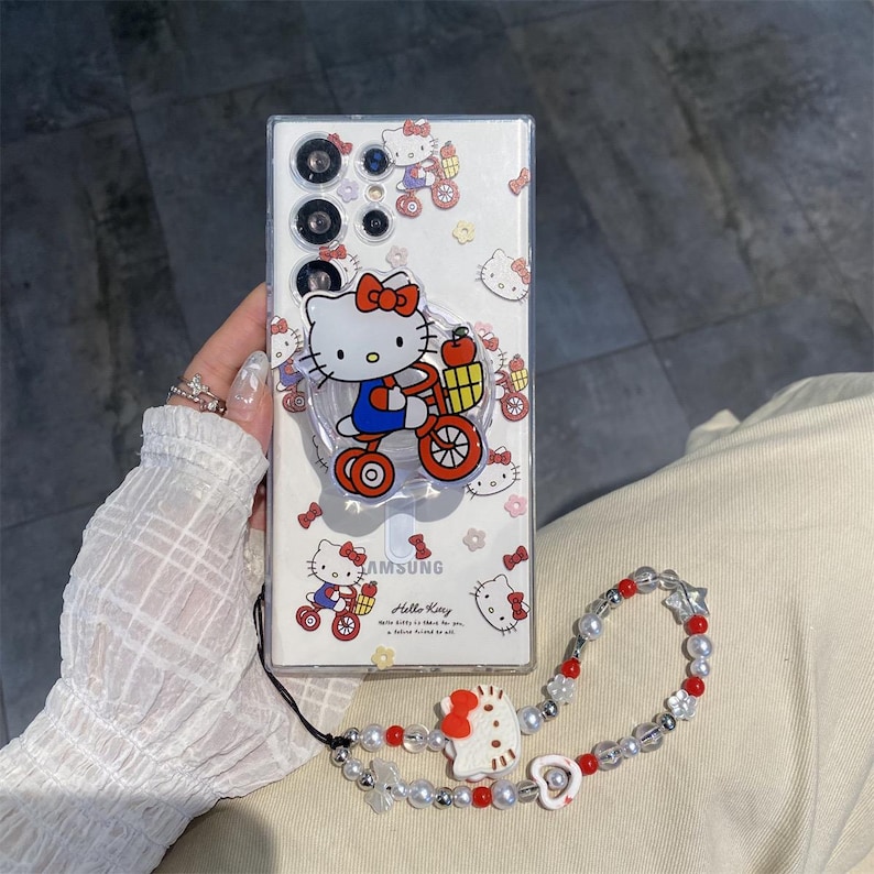 May include: Clear phone case with a Hello Kitty design. The case features a pop-up grip with a Hello Kitty graphic riding a bicycle. A matching beaded wrist strap with red and white accents is also included.
