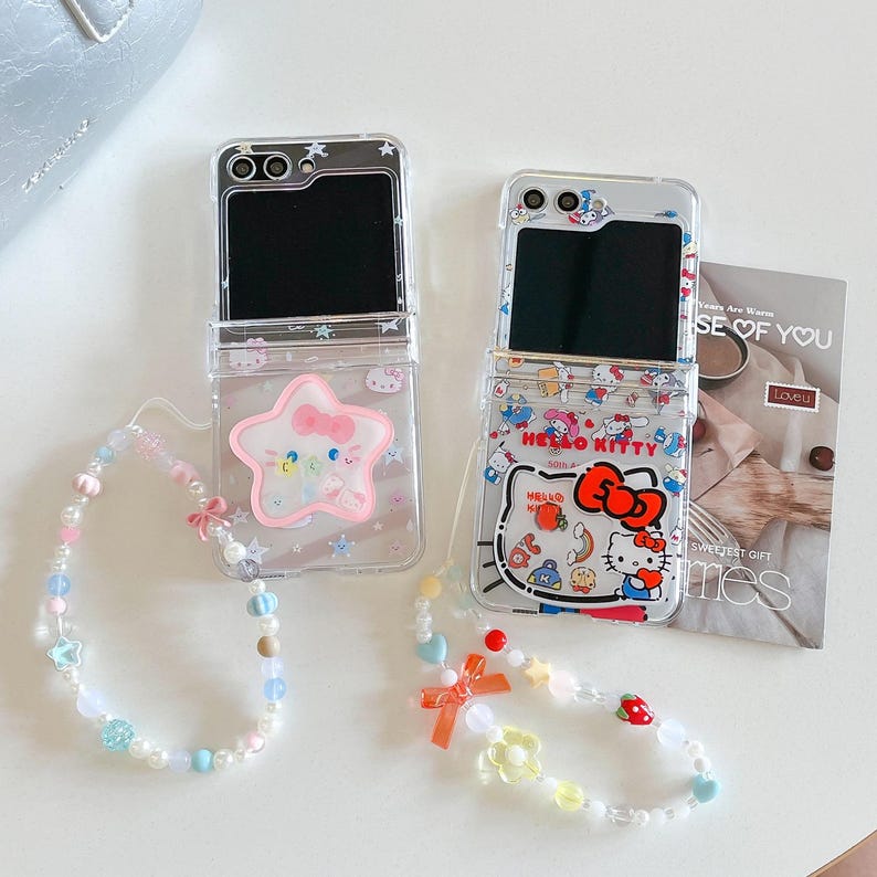 May include: Two clear phone cases with Hello Kitty designs and beaded wrist straps. One case has a star design, the other a Hello Kitty graphic. The cases are open, showing the phone screens.