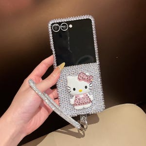 Puede incluir: Funda de tel&eacute;fono adornada con pedrer&iacute;a con un dise&ntilde;o de Hello Kitty. La funda est&aacute; cubierta de pedrer&iacute;a plateada y presenta una figura 3D de Hello Kitty con un lazo y un vestido rosas. Se adjunta una correa de mu&ntilde;eca de pedrer&iacute;a a juego.