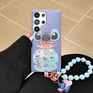 May include: A light purple phone case featuring a cartoon character face with large eyes and a wide smile. The case has a clear section with floating glitter and Stitch character designs. A blue beaded wrist strap and Stitch charm are attached.