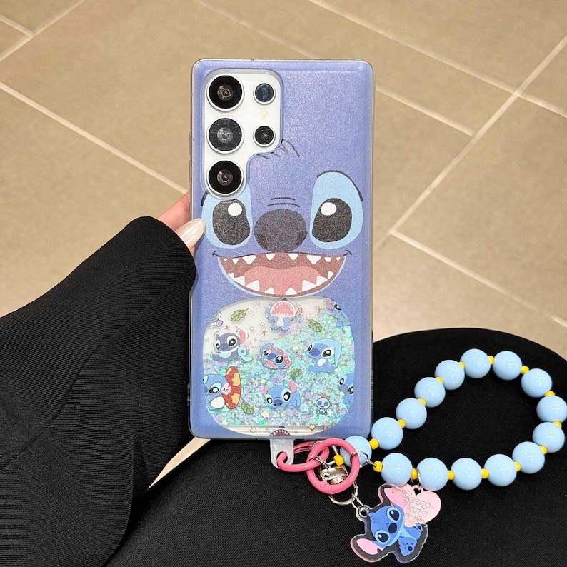 May include: A light purple phone case featuring a cartoon character face with large eyes and a wide smile. The case has a clear section with floating glitter and Stitch character designs. A blue beaded wrist strap and Stitch charm are attached.