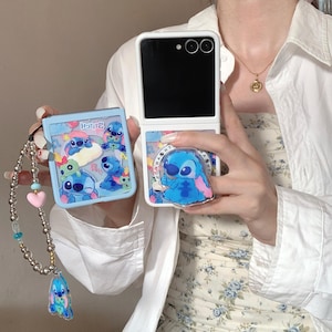 May include: A white foldable phone with a blue Stitch phone case and a matching pop socket. The phone case features various images of Stitch. A beaded charm with a Stitch charm and a pink heart is attached to the phone case.