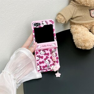 May include: Pink phone case featuring a repeating pattern of Hello Kitty faces and bows. The case has a flip design and includes a charm with a star and flower. The words "Hello Kitty" are printed on the case.