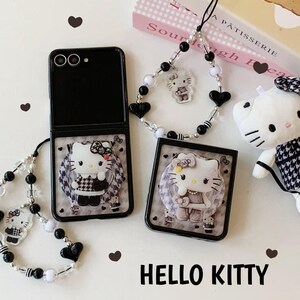 May include: Black phone case with a Hello Kitty design, featuring a black and white checkered pattern. The image includes a matching phone charm and a small plush toy, all with the Hello Kitty theme. The words "HELLO KITTY" are at the bottom.
