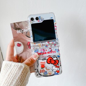 May include: Clear phone case with a Hello Kitty design. The case has a transparent design with colourful Hello Kitty illustrations, a red bow, and the text "Hello Kitty 50th Anniversary". The phone case is open, revealing the phone's screen.
